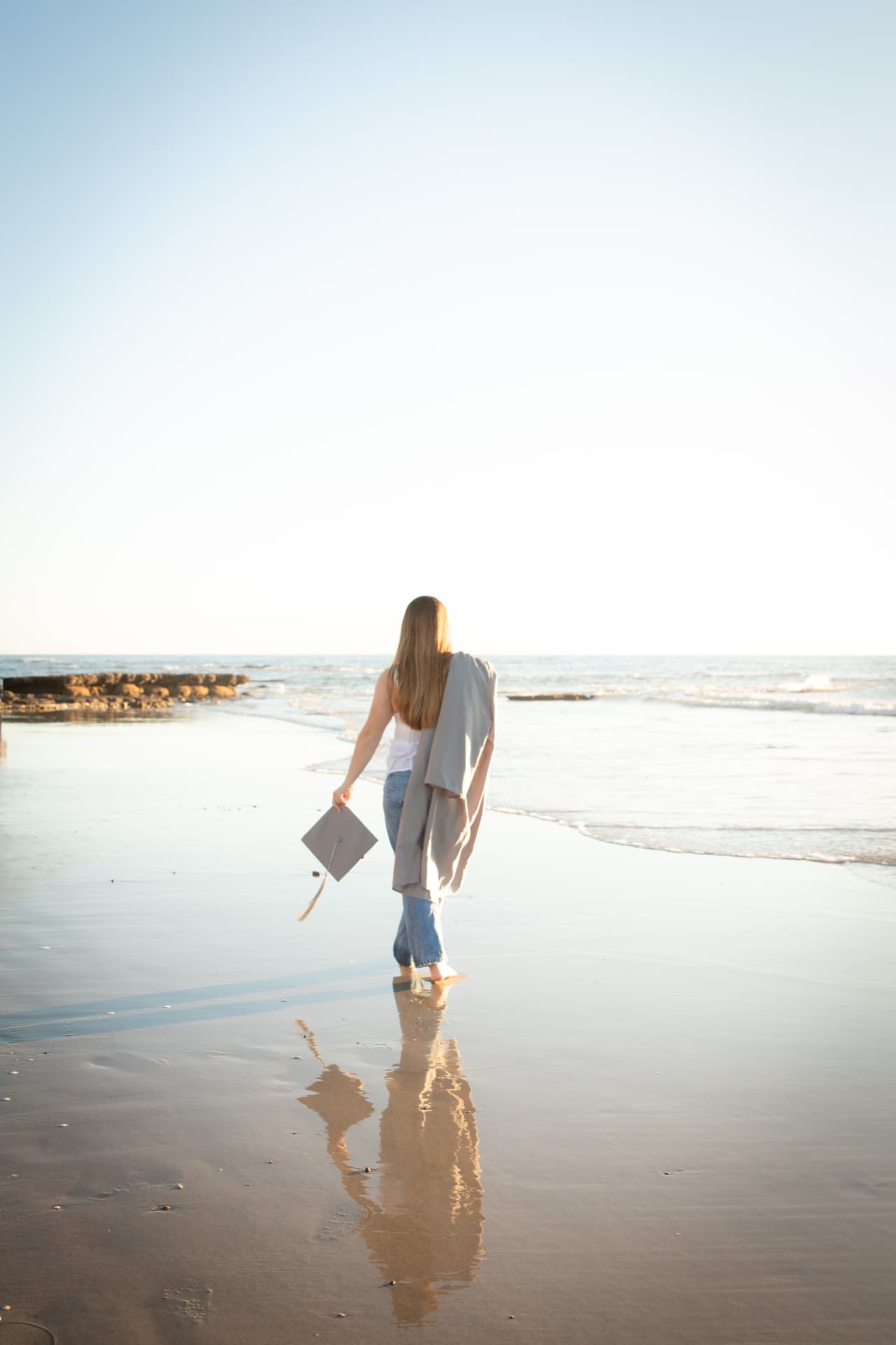 Senior walking on the wet sand holding a graduation cap at sunset