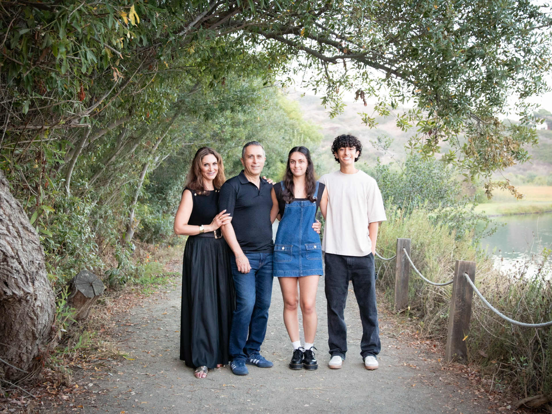 Family of four standing together on a tree-lined path by a lake