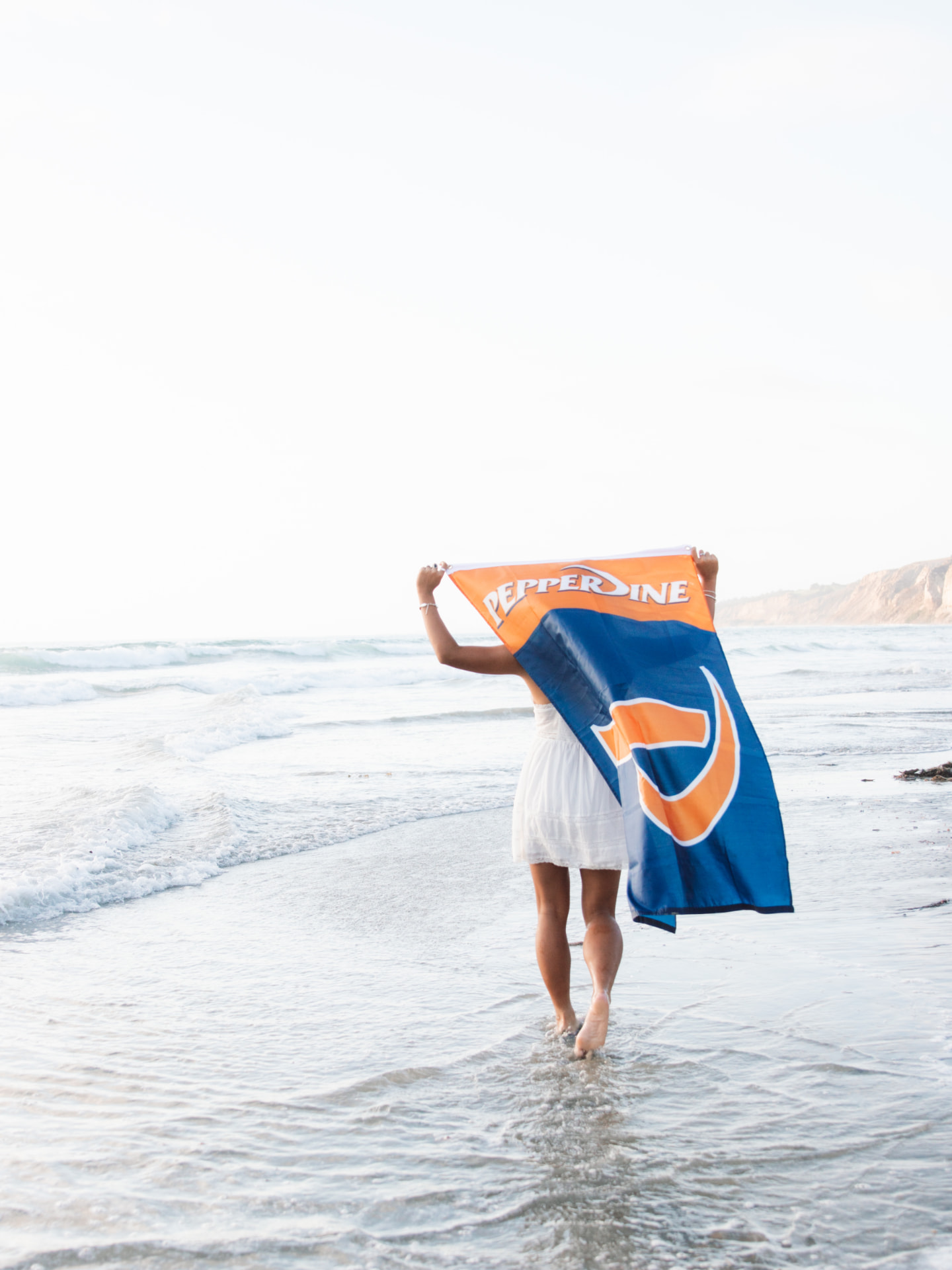 Senior walking along the shoreline holding a Pepperdine flag