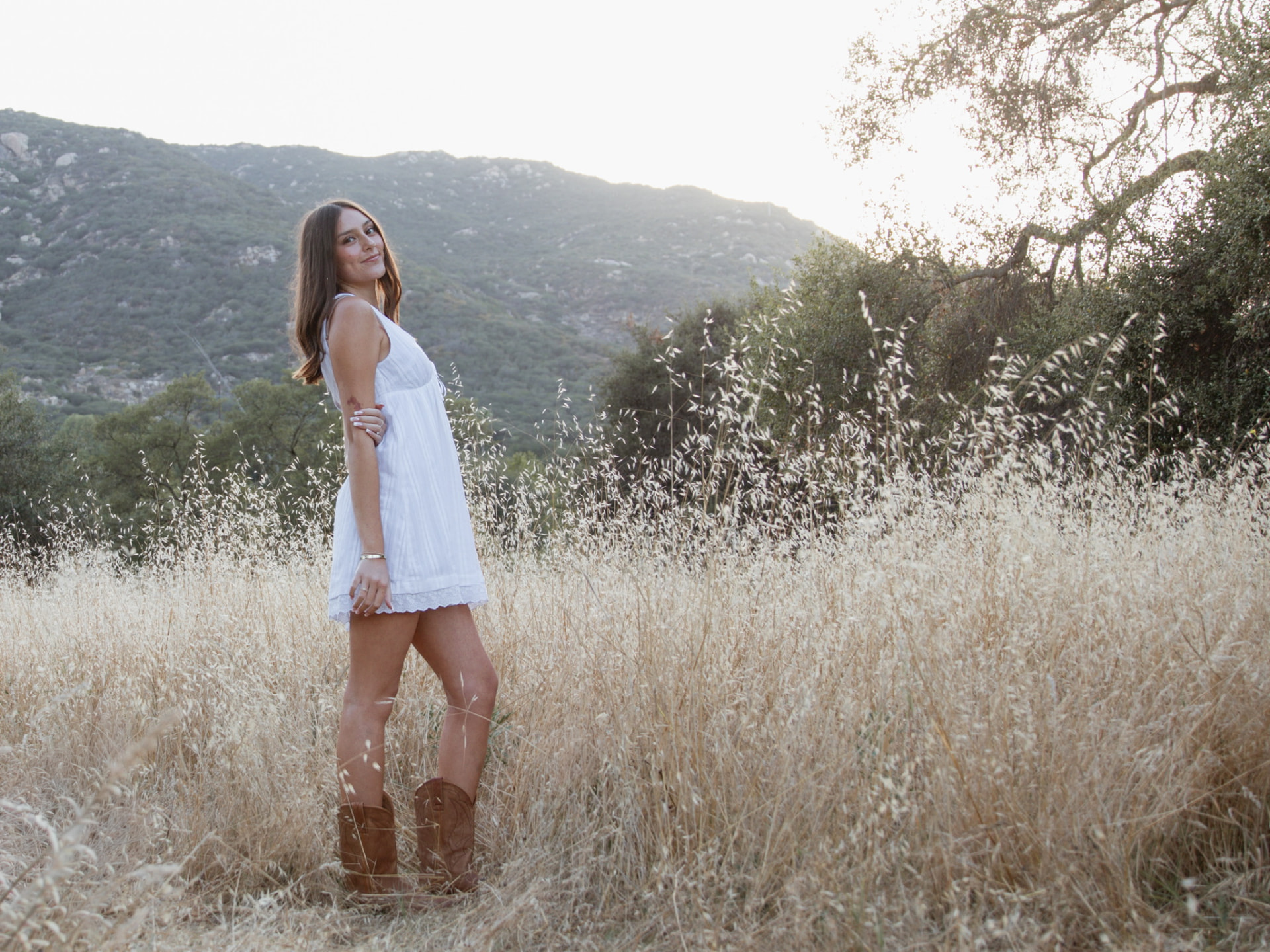 Senior in a white dress and boots standing in a golden field with rolling hills