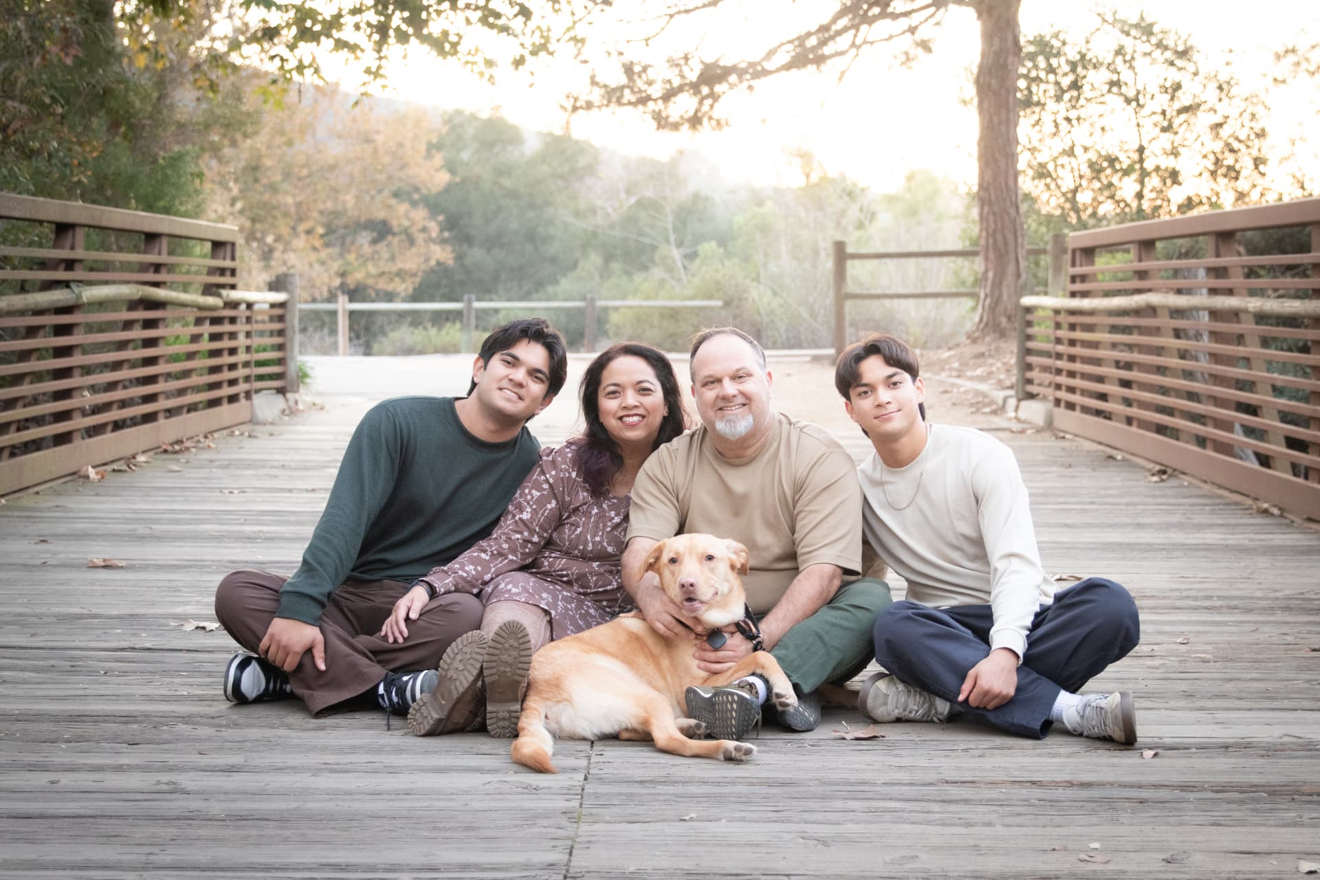Family of four and their dog sitting on a wooden bridge at sunset