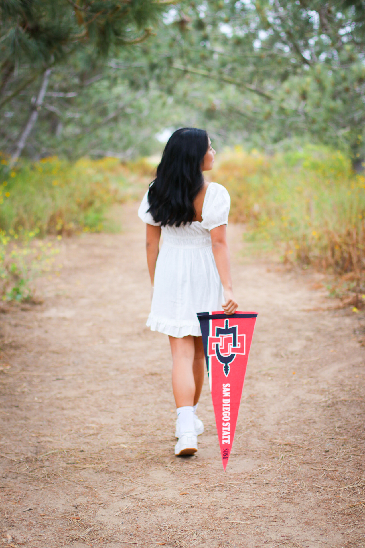 Senior in a white dress walking down a path holding a San Diego State pennant
